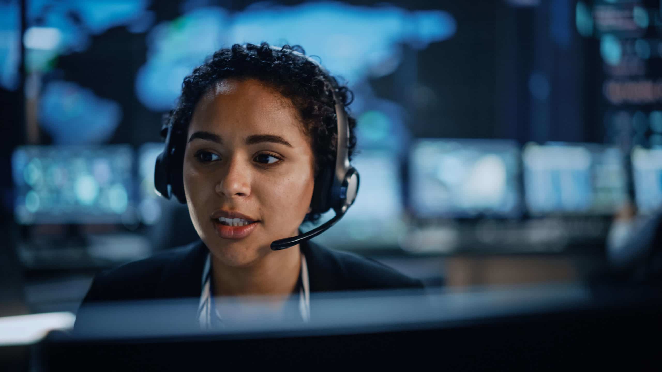 Portrait of Professional Technical Support Female Specialist Working on Computer in Monitoring Control Room with Digital Screens. Employee Wears Headphones with Mic and Talking on a Call.